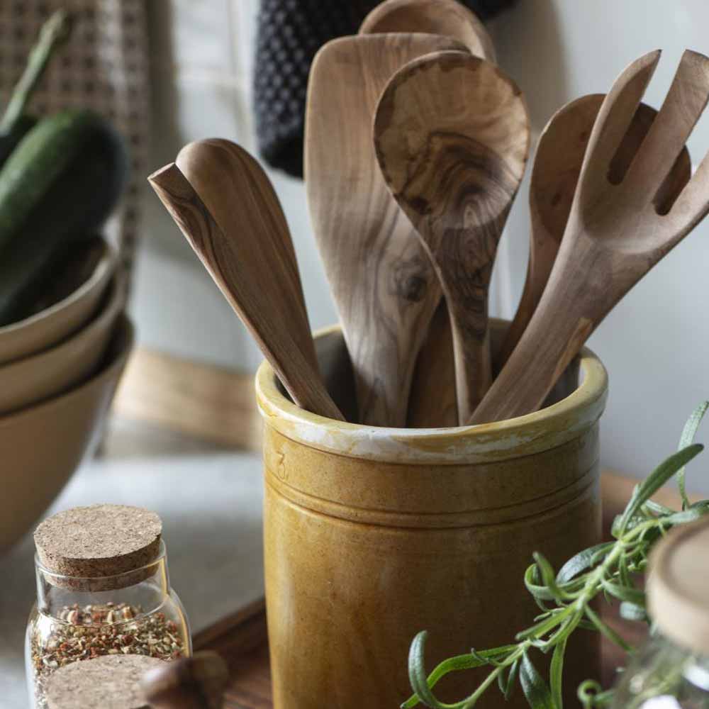 Verschiedene gemaserte Holzlöffel und ein Salatbesteck stehen in einem olivbraunen Gefäß neben einem Holzbrett