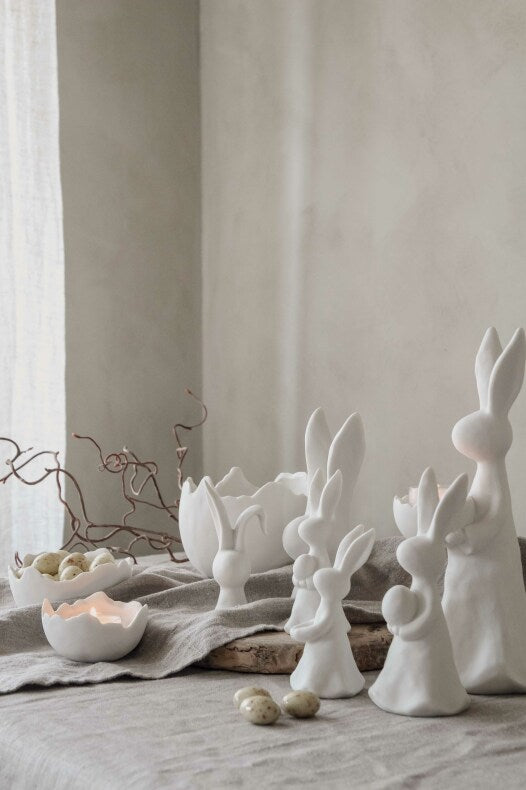 Eine Sammlung von Majas Cottage – Hase groß, ausgestellt auf einem strukturierten Tuch in der Nähe einer brennenden Kerze.