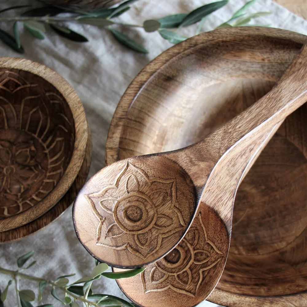 Nahaufnahme des aus Holz geschnitzten Majas Cottage – Salatbesteck Fleur 2er-Sets mit Löffeln und Schüsseln mit Blumenmuster, platziert auf einem mit grünen Blättern geschmückten Tisch.