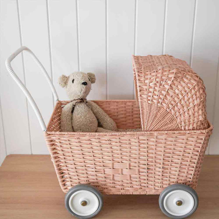 Ein Olli Ella - Puppenwagen Strolley rosa, der in einem Korbkinderwagen mit weißen Rädern vor einer weiß getäfelten Wand sitzt.