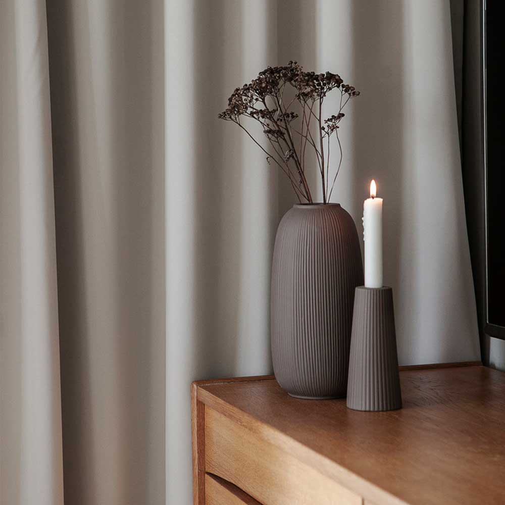 Eine Storefactory – Åby Vase Keramik braun mit getrockneten Blumen und einer brennenden Kerze auf einer Holzoberfläche vor einem vorgehängten Hintergrund.
