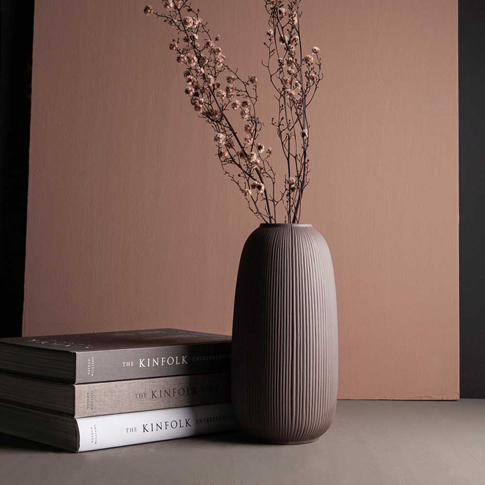 Braune gerillte Vase mit trockenen Zweigen neben gestapelten Büchern vor einer rostbraunen Fläche