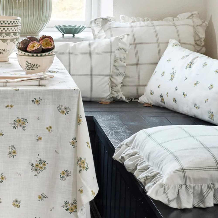Eine gemütliche Ecke mit einer Kollektion von Strömshaga - Kissenbezug Alma 45 x 45 cm Kissen und einer geblümten Tischdecke auf einer Holzbank.